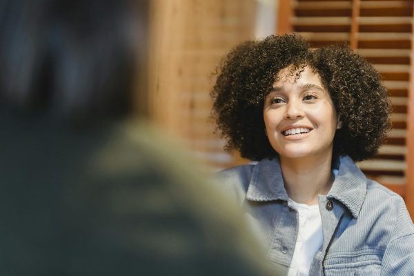A cheerful young woman with curly hair smiling during a casual indoor conversation.